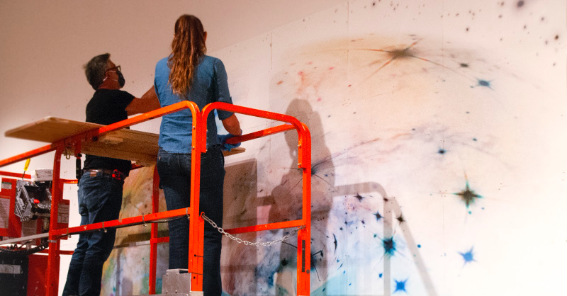 SDMA's tweet photo. This huge galaxy #drawing is one of 2! 💫
Here, our Assistant #Registrar closely examines the condition of each paper panel before handing them off to our Chief #Preparator and #MuseumTechnician for #installation—using magnets and a carefully laid out grid on our #gallery wall.