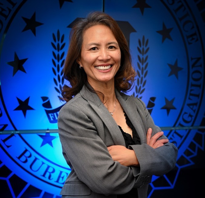 a smiling business woman of Asian descent with her arms folded standing in front of a backlit blue FBI seal. 