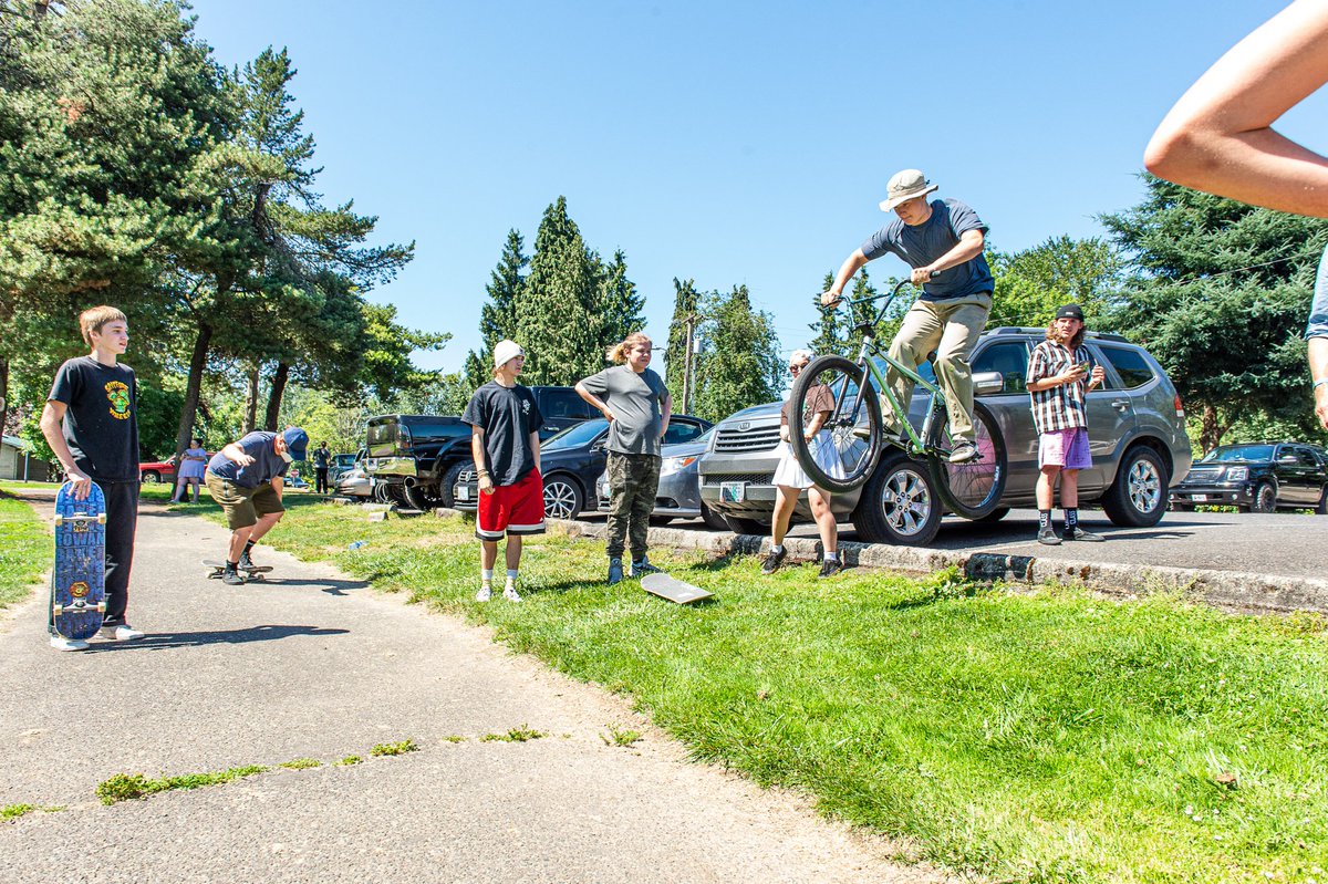 A teenage boy jumping a length of grass been a parking lot and walkway on his BMX bicycle. Six friends stand by watching. It is very sunny and rad as hell.