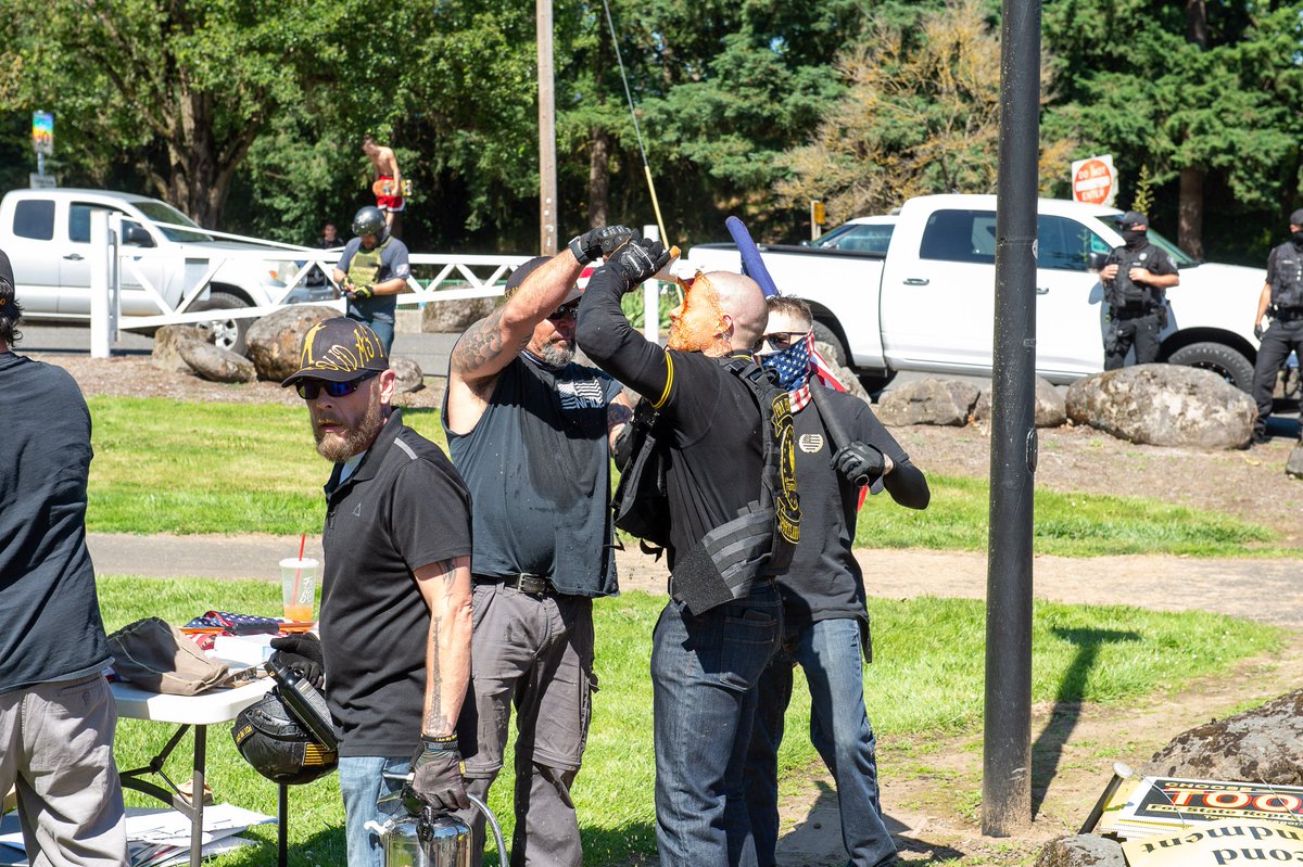 Four white grown men standing around in black and yellow tactical clothing. One man appears to be pouring a paper cup of cola onto another man's face. A third man stands by holding a US flag over his shoulder.