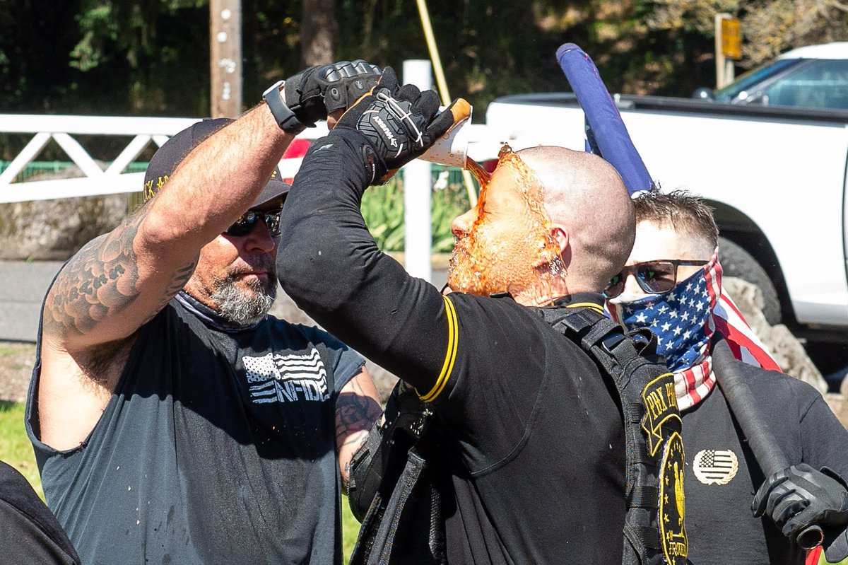 Even closer cropped frame. This time it is very clear that it is indeed an at least 20 ounce cup of cola beyond dumped directly on a bald man's face by his friend, who is wearing a black shirt with a black and white American flag with the word 'infidel' inside the stripes.