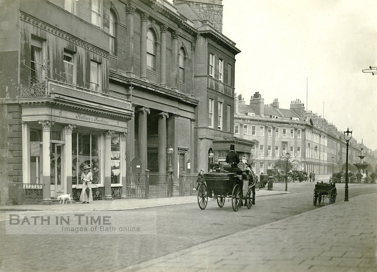 Argyle St c. 1910. A lady watches a horse drawn carriage pass by.  Behind her a hat shop and the Argyle Congregational Chapel.  In the background horse drawn taxis can be seen waiting in Laura Place.