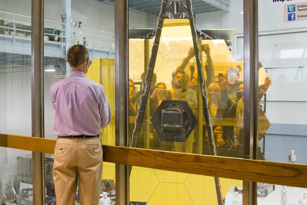 John Mather, Webb's Senior Project Scientist, faces the James Webb Space Telescope primary mirror, taking a selfie. About a dozen people standing behind Dr. Mather are visibly reflected in the mirror. This image was taken at NASA's Goddard Space Flight Center in May 2016.