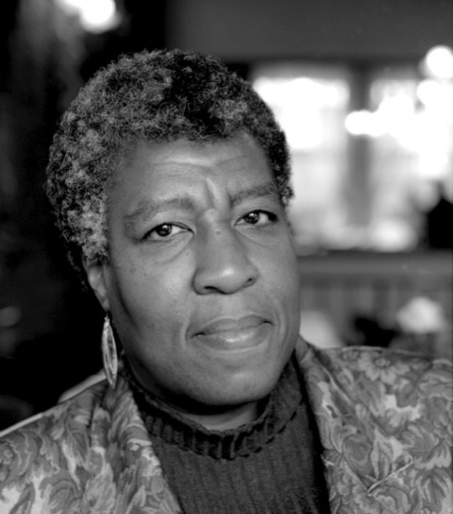 Octavia E. Butler in black and white film. She is in focus with a blurred background and looks thoughfully into the camera.