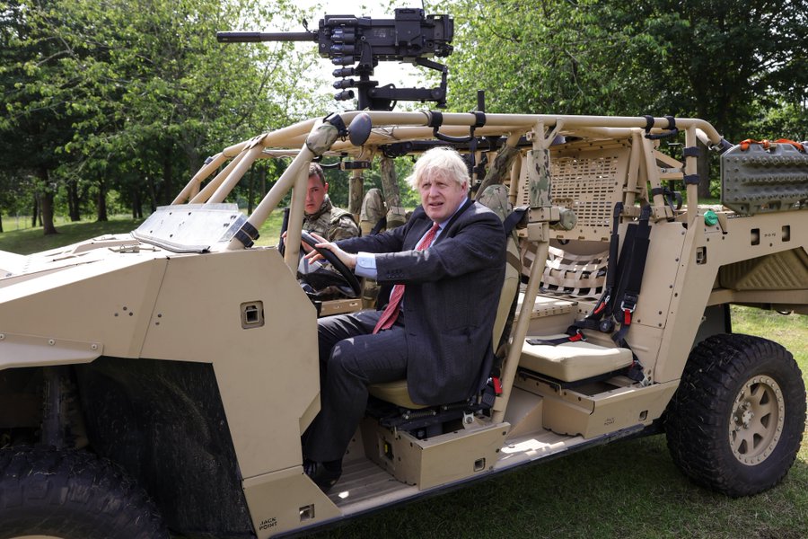 Boris Jonson sat in a military vehicle driver seat. He is wearing a suit. The vehicle is desert colour and has a machine gun on the tip. It is parked on grass.