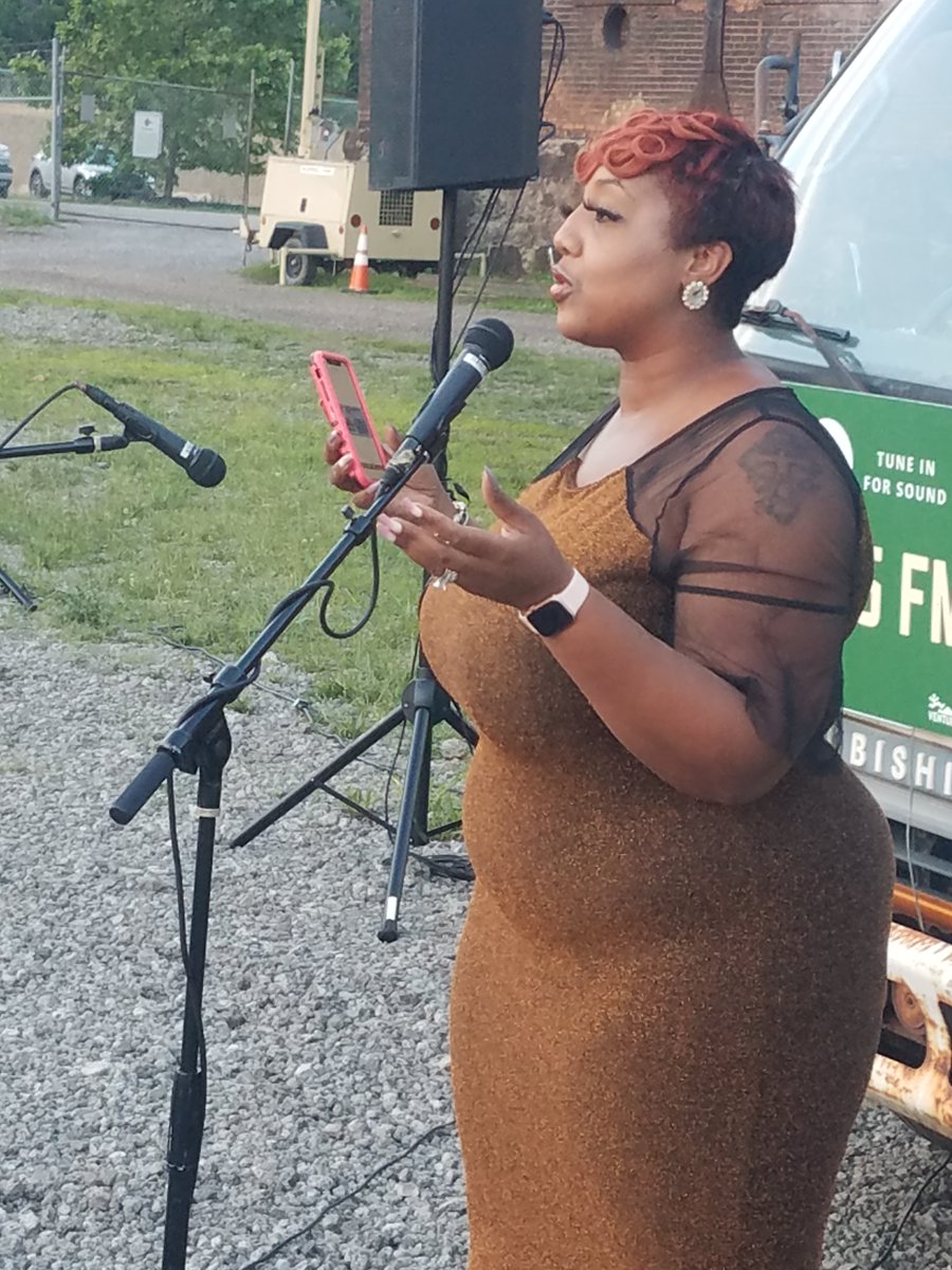 Merecedes J Williams of Movie Scene Queen stands in front of a microphone speaking to an audience (not pictured). Merecedes' hair is reddish and she wears a velvet sand-colored dress. She stands on the gravel ground.