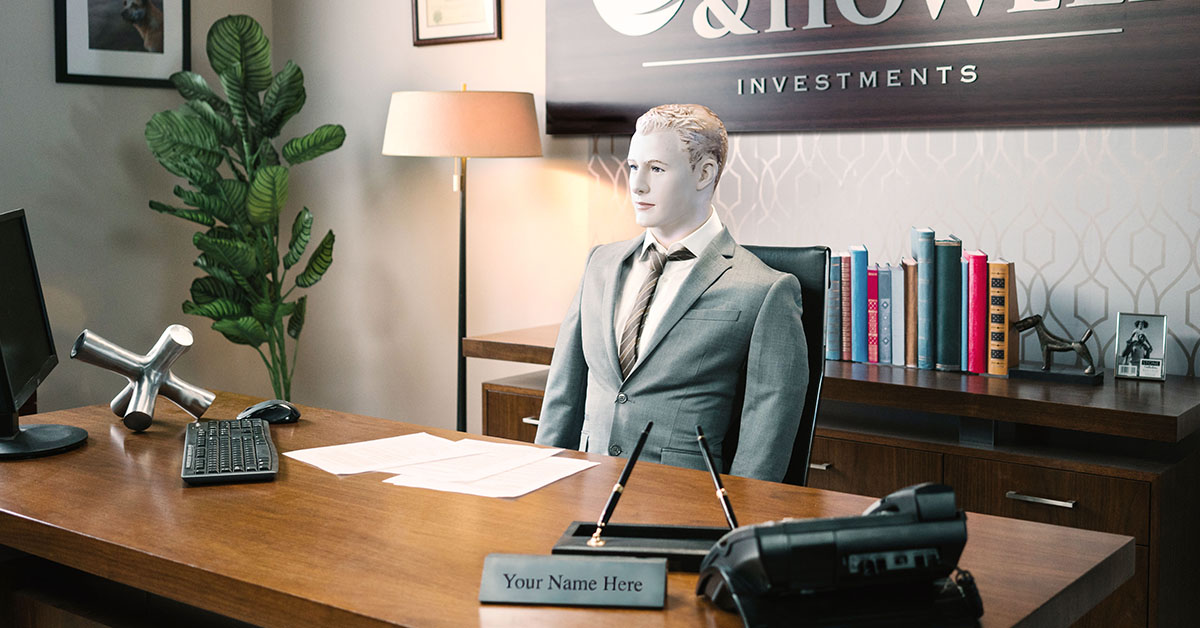 Mannequin in a gray suit is seated in an office behind a desk with a nameplate reading “Your Name Here” 