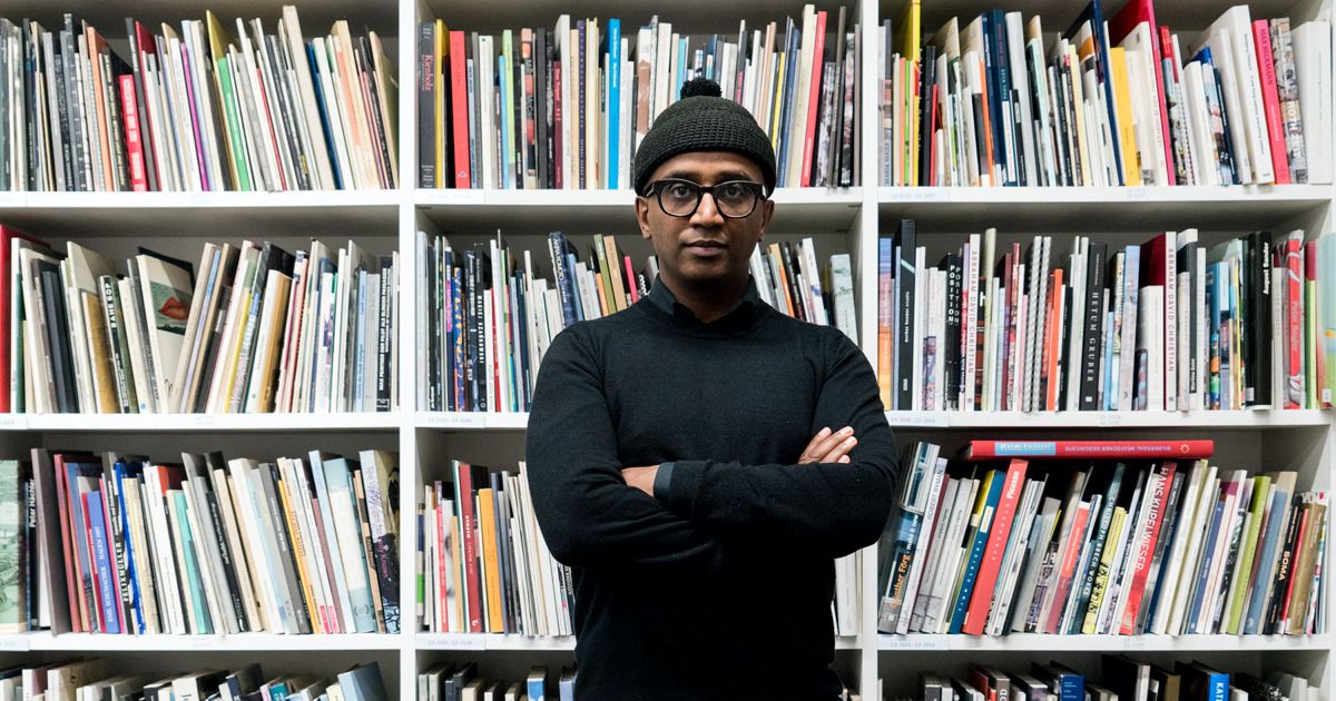 Un homme portant des lunettes, une tuque et un chandail à manches longues noires ainsi que des pantalons noir et blanc et des chaussures blanches se tient debout devant une étagère de bibliothèque remplie de livres.