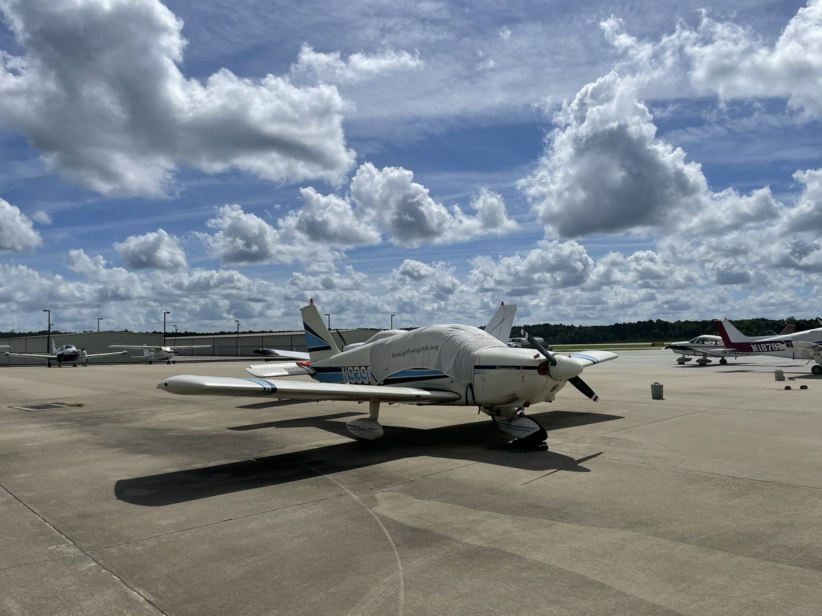 A low-wing single-engine aircraft with a cover over the windows. The cover says “RaleighFlyingClub.org”. The plane is parked on a tarmac, with lots of other small airplanes parked nearby. The sky is spotted with fluffy clouds, with mostly blue sky above.