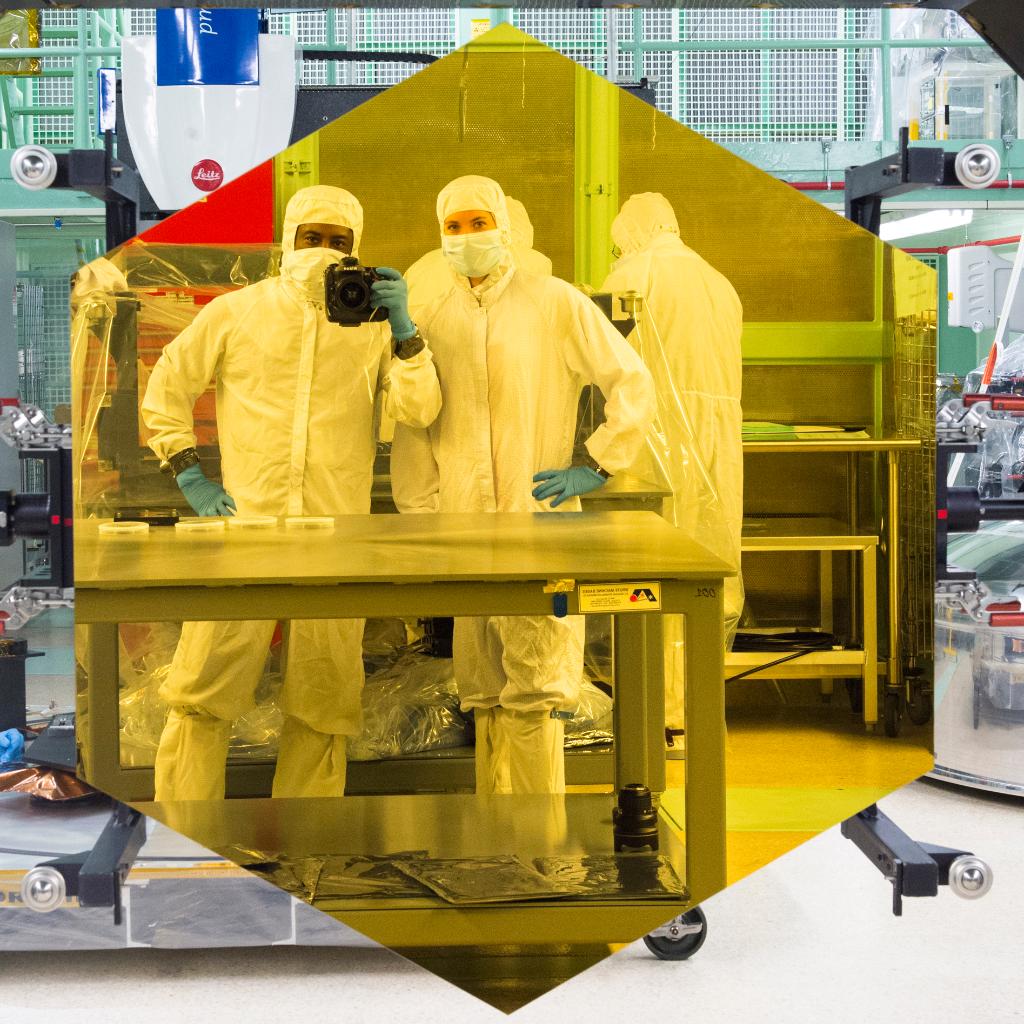 Webb team photographers Chris Gunn and Desiree Stover take a selfie in front of a gold-coated, hexagonal Webb mirror segment. Both are in cleanroom suits. Chris Gunn is holding the camera in one hand.