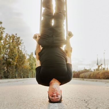 Un jeune homme se tient en équilibre sur sa tête au centre d'une route longée d'arbres. Il tient des béquilles dans ses mains.