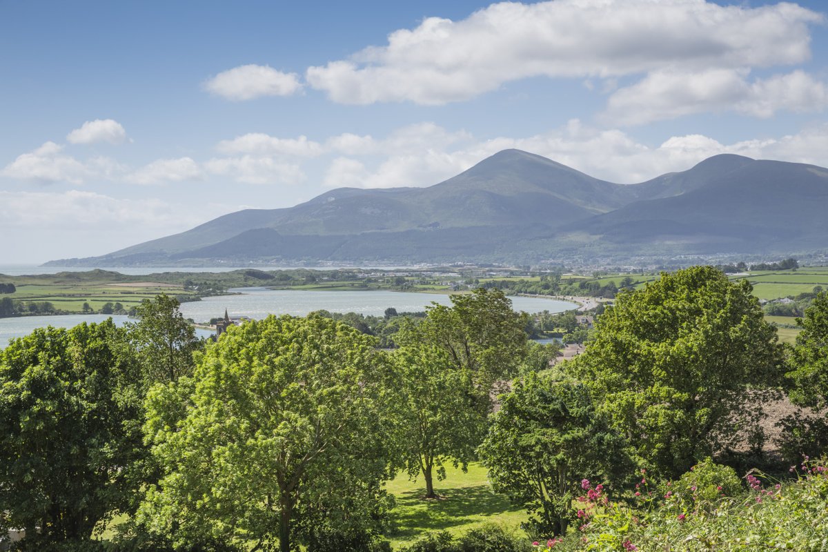 An orchard of trees in the foreground, in front of an inlet from the sea. In the distance, mountains rise up along the horizon, and white clouds float through a bright, blue sky.