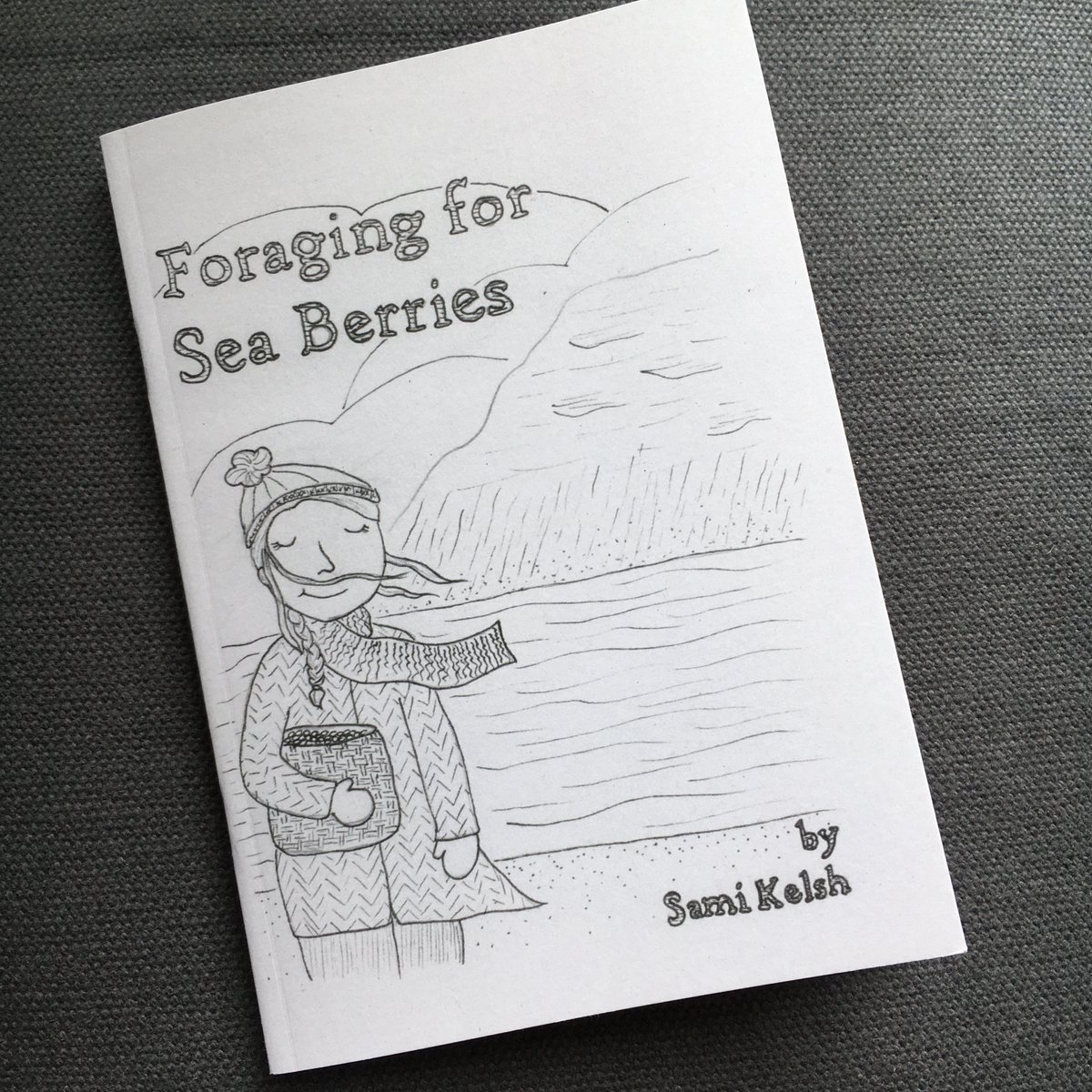 The cover of the book Foraging for Sea Berries by Sami Kelsh. A white woman stands on the shore of a cold sea. She is wearing her hair in a side braid with a knitted hat with a pom pom, a long scarf blowing in the breeze, and a thick wool coat. She is holding a basket of berries.