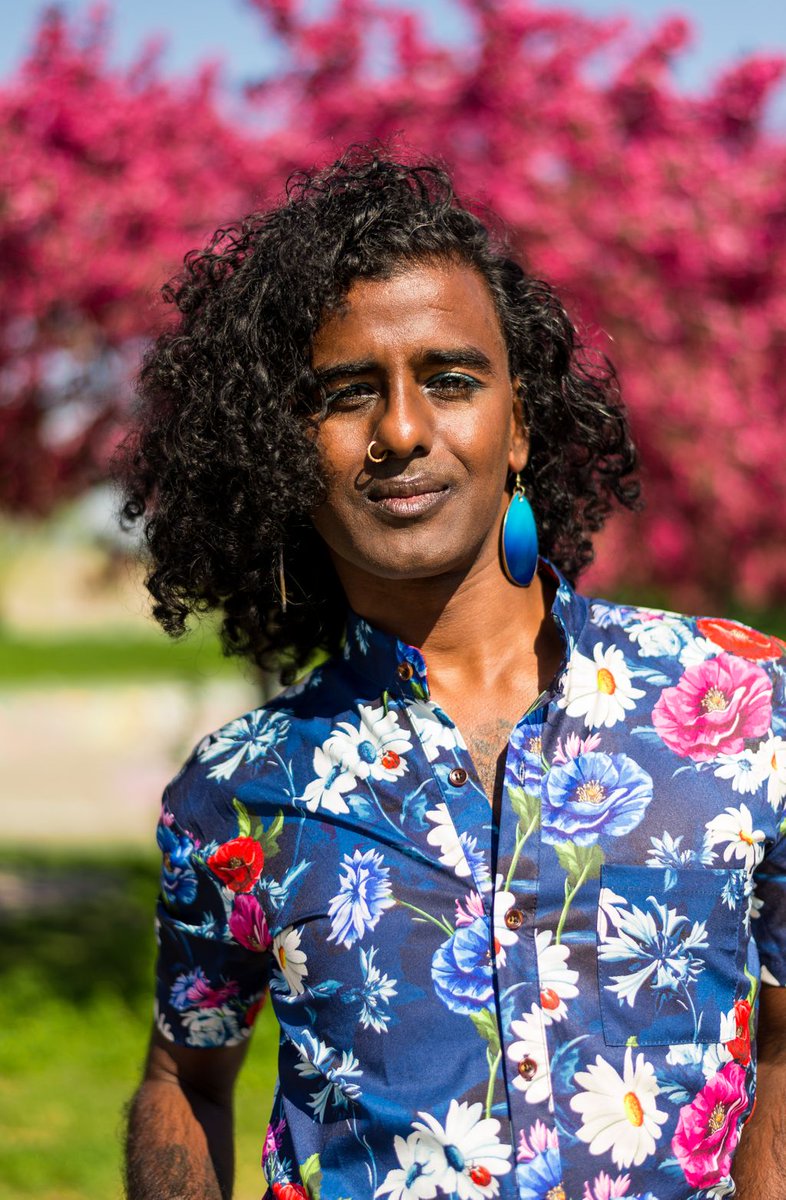 Un.e artiste noir.e aux cheveux noirs bouclés se tient debout dehors. Iel porte une chemise à manche courte colorée et fleurie.  