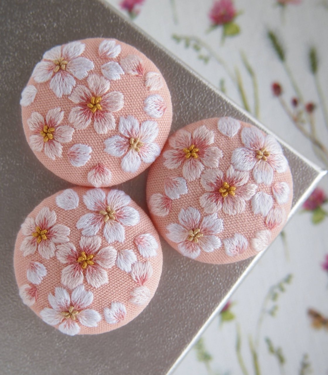 ララの花かんむり 刺繍で楽しむ花の物語 発売 満開の桜を眺めるほんのひとときの胸のときめきを表現 しました 優しいサーモンピンクの地にぽっと色づく桜の花びらを刺繍しています 直径3cmの刺繍部分にぷっくりとした花をぎゅっと詰め込んだ華やかな