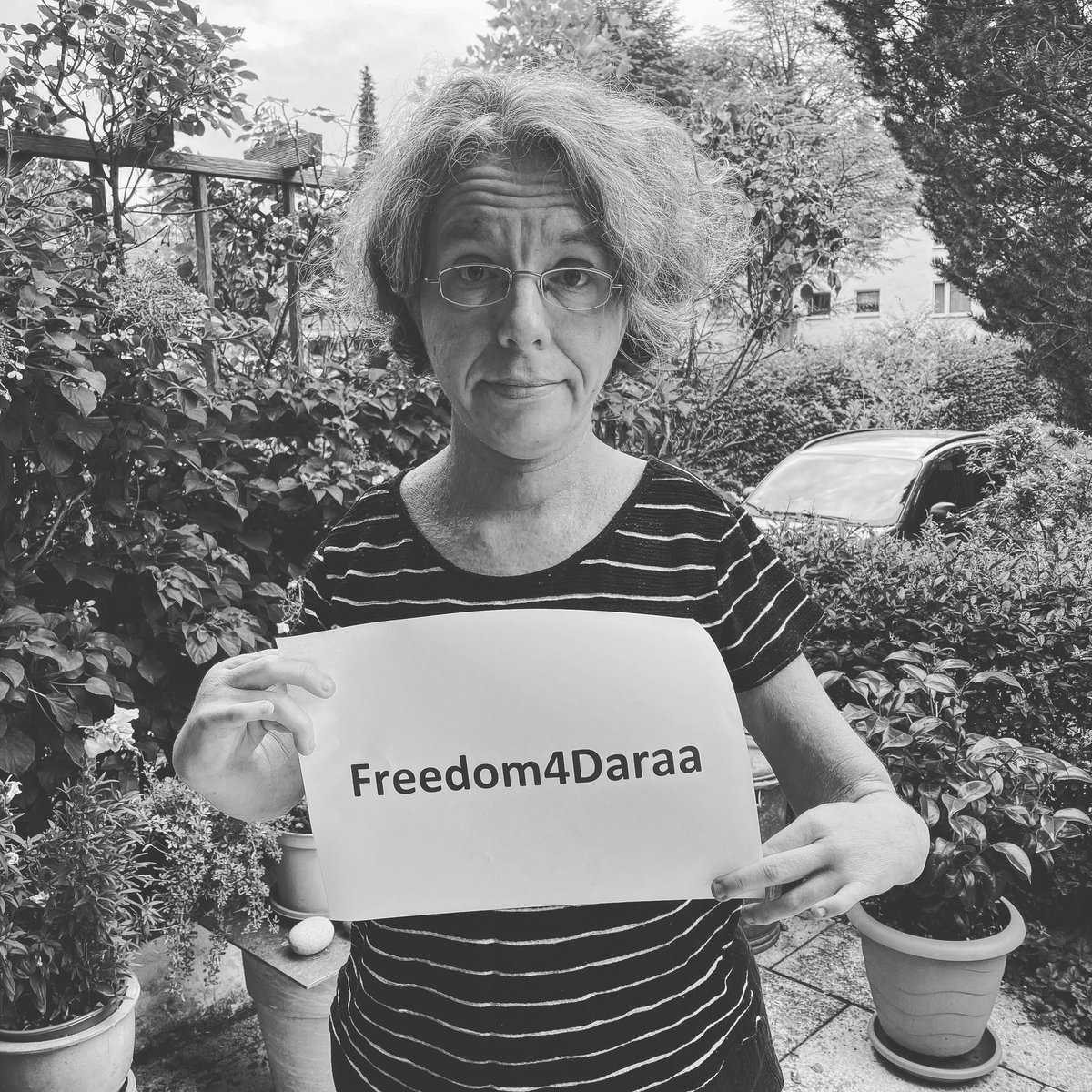 Schwarzweiß Foto. Kathrin steht auf der Terrasse und hält ein Papierschild in den Händen. Aufschrift:Freedom4Daraa