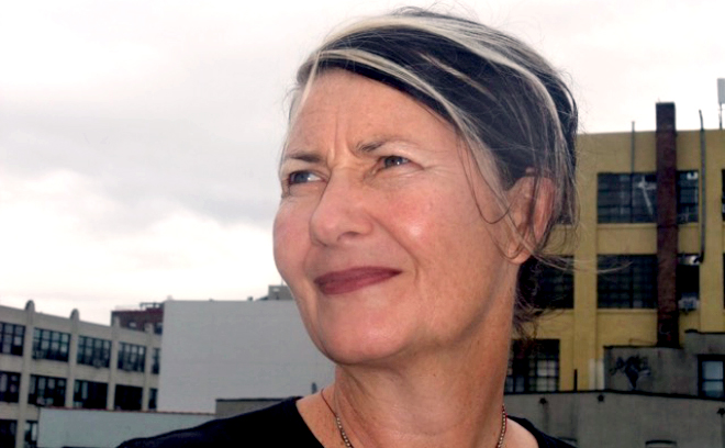 Designer Marlene McCarty, a woman with streaks of dark and light hair pulled back into a ponytail, looks off to the side in front of a few industrial-looking buildings