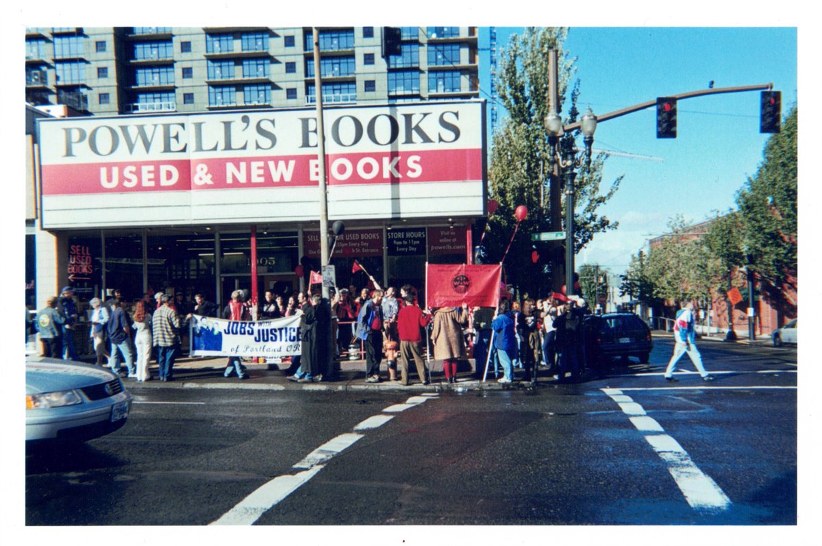 2003 photo of the 10th and Burnside entrance to Powell's City of Books in Portland, Oregon. A crowd is gathered on the sidewalk under the marquee, carrying flags, balloons, and banners. A banner on the left reads, "Jobs with Justice of Portland, OR," and a banner on the right reads, "I.W.W."