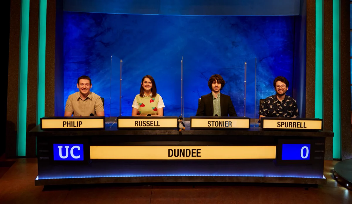 The University of Dundee's 2021 University Challenge team in the studio. The four participants are sitting facing the camera behind a desk with Dundee on a lightbox, and each of them have a lightbox with their surnames - Philip, Russel, Stonier, Spurrell - in front of them.