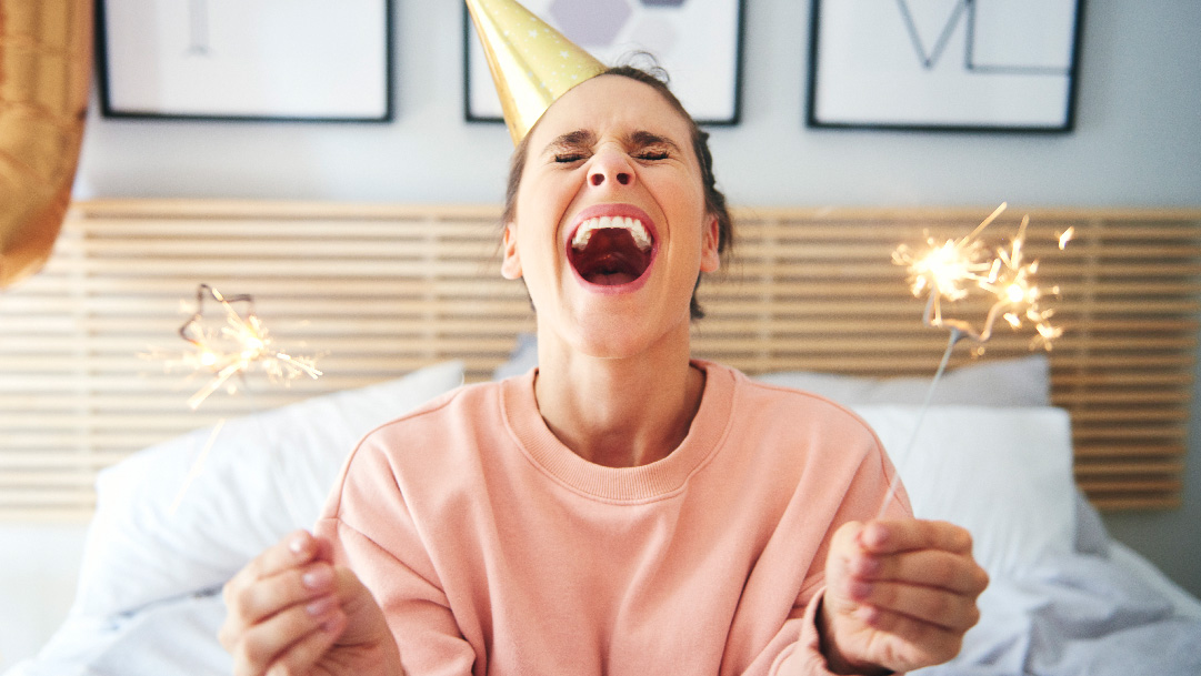 Woman in gold party hat laughs while holding two sparklers 