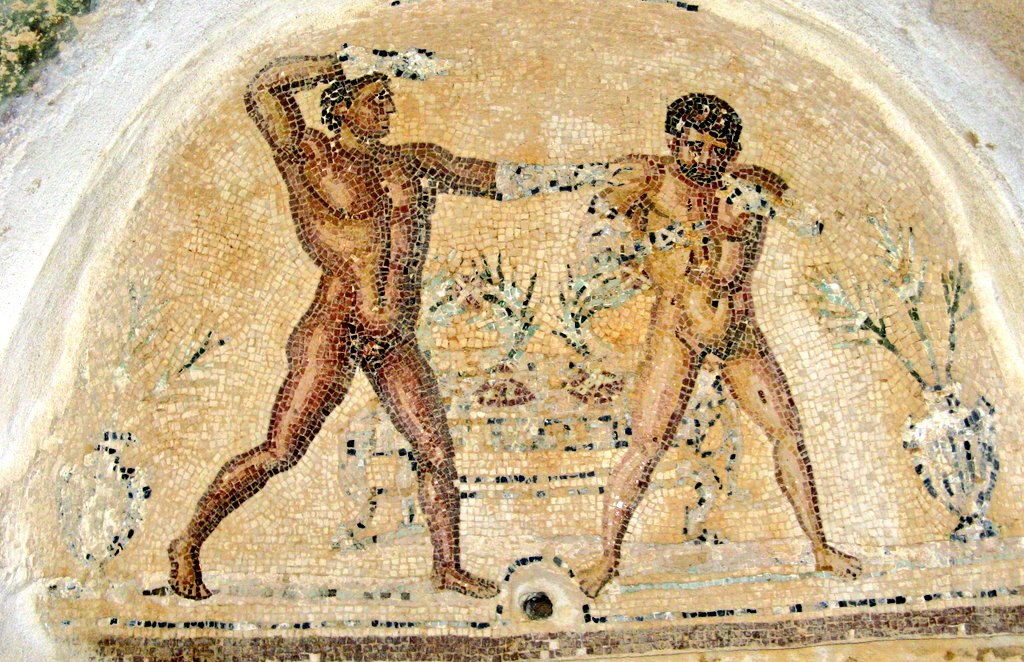 Two naked, muscular men stand in front of a table on which stand two victory wreaths boxers. Either side of them are silvery looking amphorae, each holding three palm fronds, also prizes for victors. The man on the left is turned towards his rival, his right fist raised threateningly, his left hand on his rival's shoulder. The man on the right holds his arms behind his back, and his head is bowed in submission. The victor has darker skin and is beardless, so presumably younger; the loser has a beard. The victor's arms and hands probably were clad in Roman boxing gloves - leather strapping often reinforced with metal - but those parts of the mosaic are damaged. The mosaic is in the arch of an apse; a hole in the centre at the bottom, between the two boxers' feet, suggests a spout poured water, perhaps into a bowl on a pedestal in the alcove.