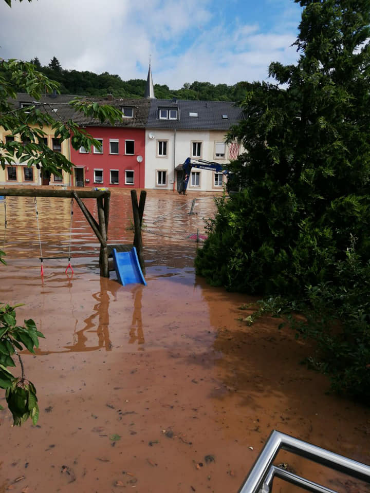 Überfluteter Spielplatz und Straße in Ehrang. Im Hintergrund Häuser, bei denen das Wasser fast bis zu den unteren Fenstern steht.