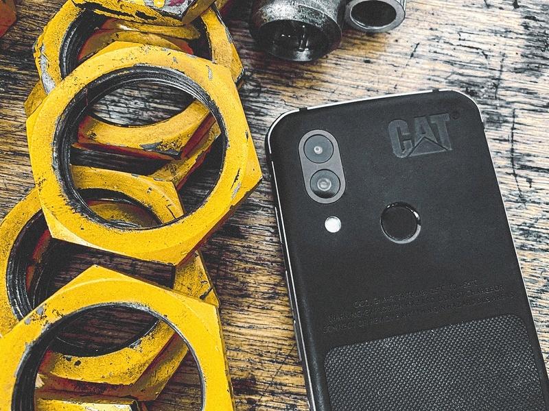 A rugged Cat S62 smartphone sits on a wooden workbench with yellow steel rings next to it.