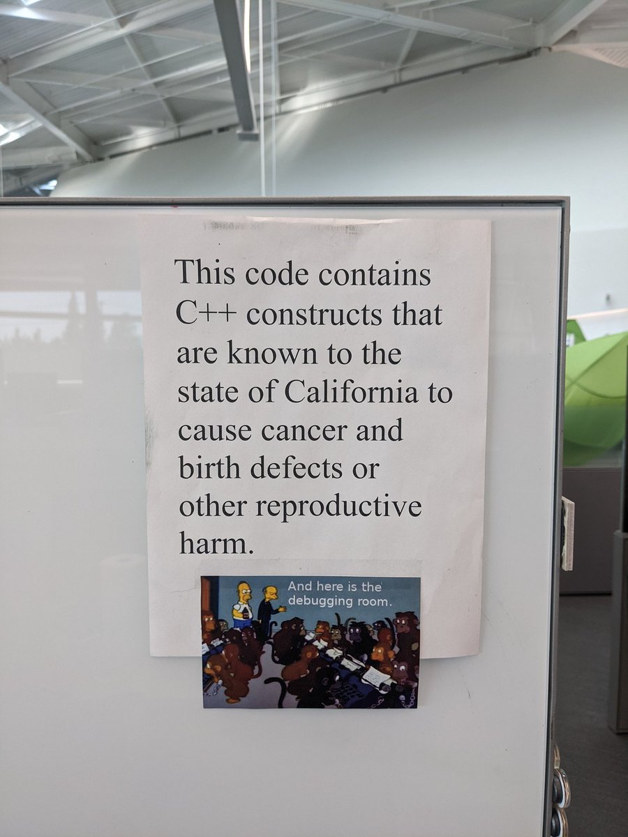 A paper sign on a desk that says "This code contains C++ constructs that are known to the state of California to cause cancer and birth defects or other reproductive harm". Below it is a Simpsons meme where Mr Burns shows Homer a room full of monkeys, with the caption reading "and here is the debugging room".