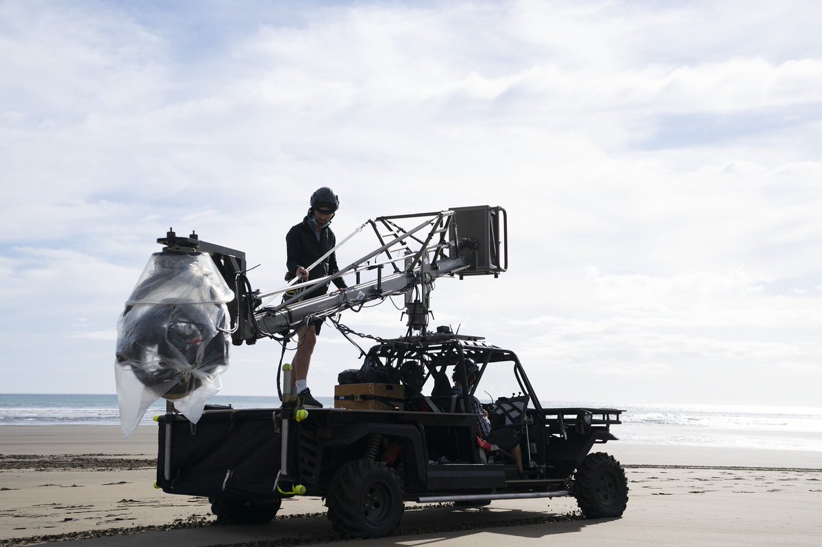 A crew member stands on the back of a camera truck on a sandy beach guiding a jib/crane.