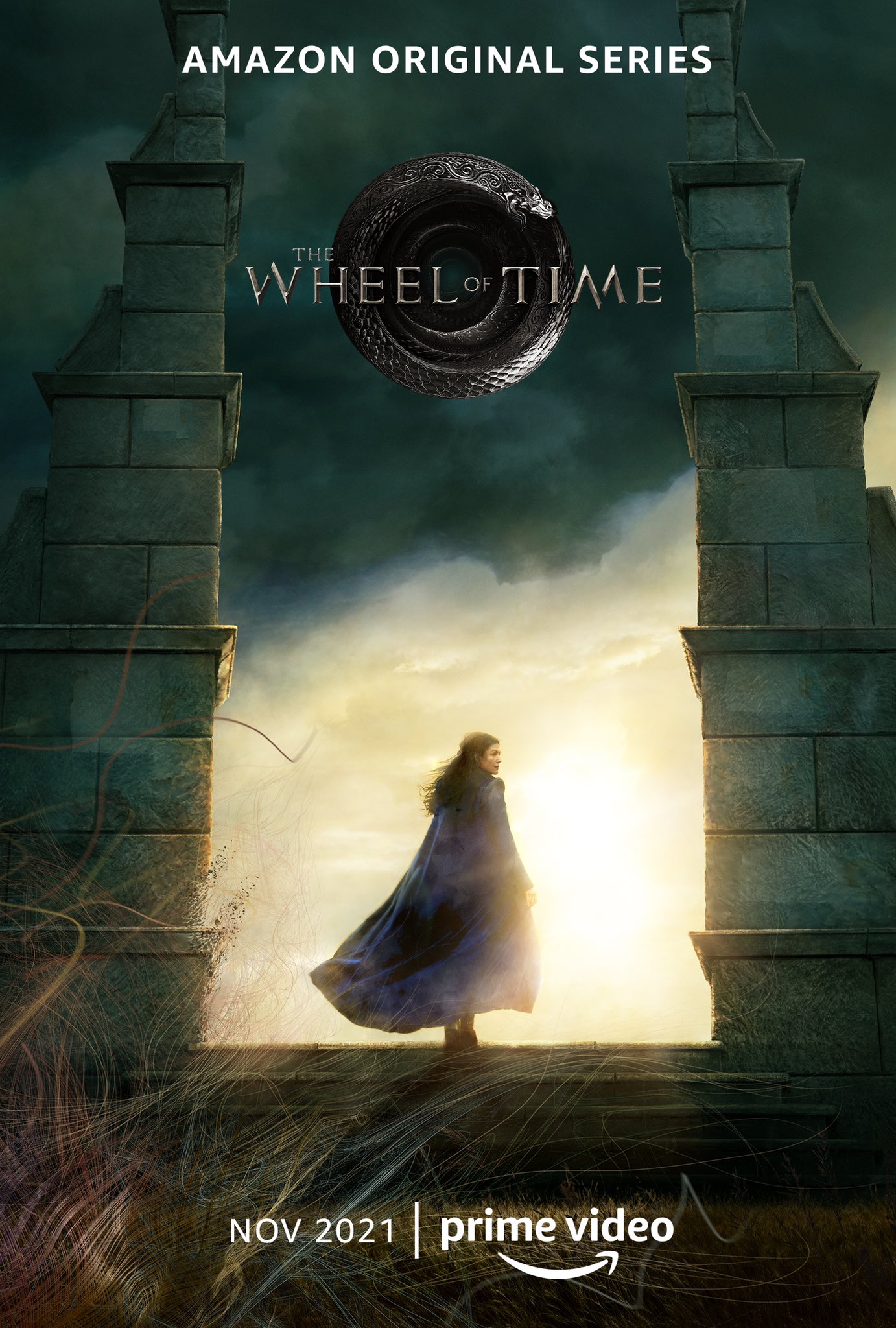 The poster depicts an iconic moment from The Eye of the World. Moiraine, standing beneath slowly moving clouds and wrapped in a blue cloak that is blowing in the wind with its hood down, looks over her shoulder at the audience as she steps through a Waygate into the unknown. She is inviting us to go on a journey with her. The words "Amazon Prime Original" and "Nov 2021" are overlaid on the image along with the Wheel of Time serpent logo and the Prime Video smile.