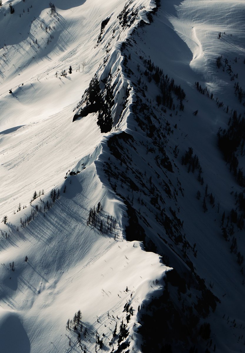 An aerial photo of a snowy mountain chain. The sun is illuminating the left side of the mountain tops and creating dark shadows on the right side.