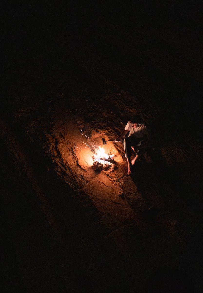 An aerial photo of a person sitting next to a small bonfire. Outside the glow of the fire, the frame is entirely black.
