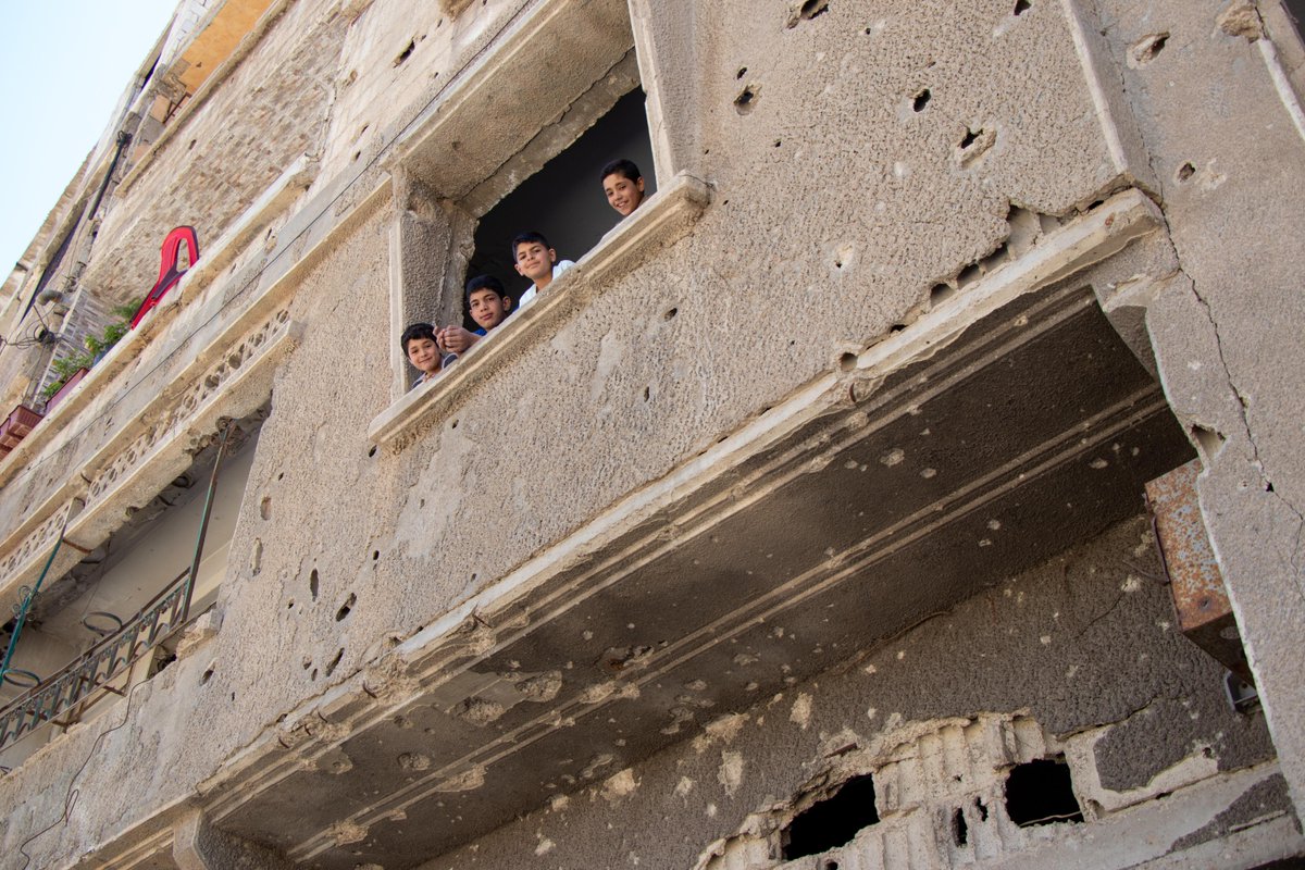 Quatro crianças olham para a rua debruçadas em uma janela de um edifício danificado e com marcas de balas na Síria.