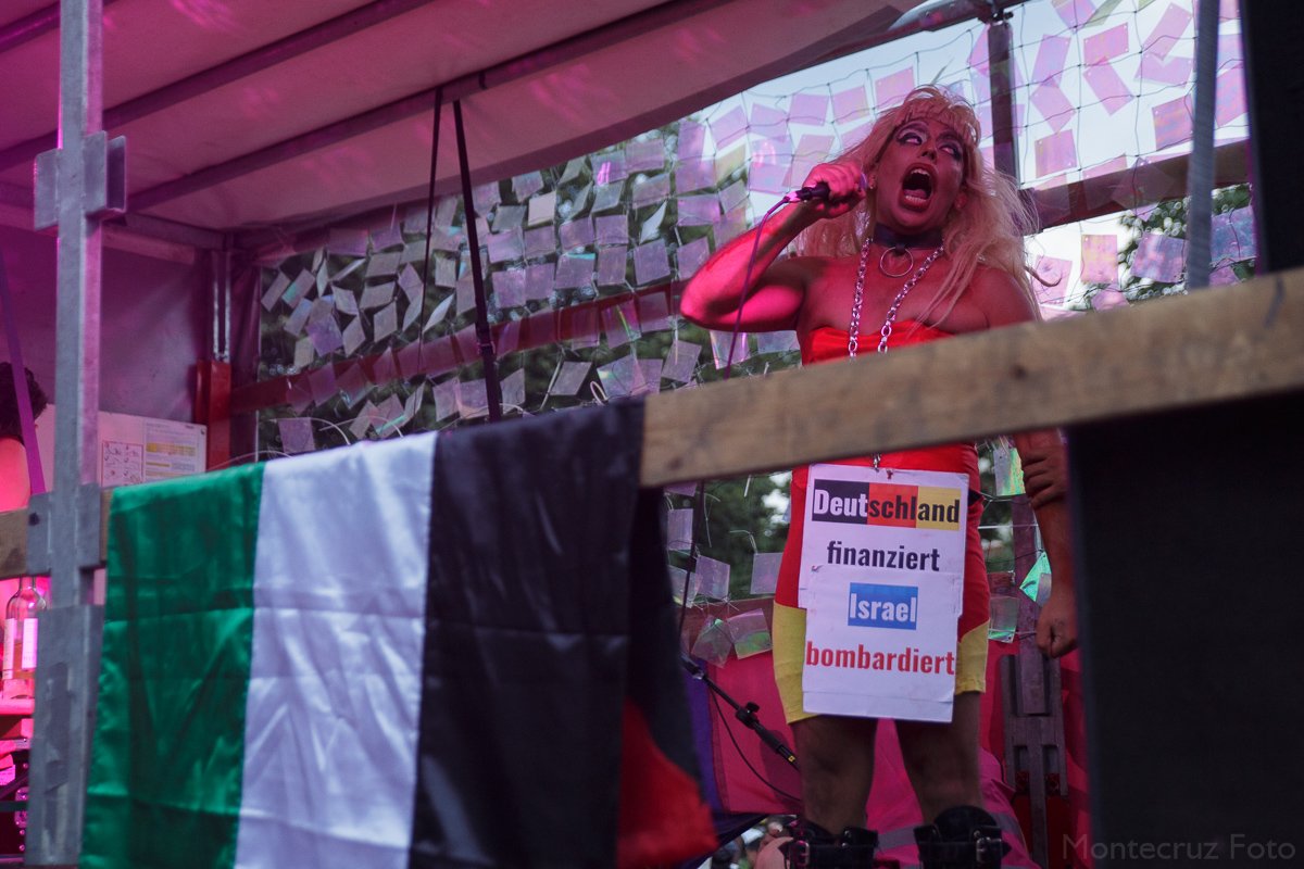 Im Bild: Redner*in bei Queer Pride mit Statement "Deutschland finanziert, Israel bombardiert"