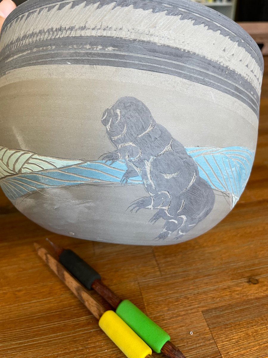 Side view of a cute little black tardigrade against a backdrop of blue stylized water on an unfired pot about 6 inches high and 8 inches across