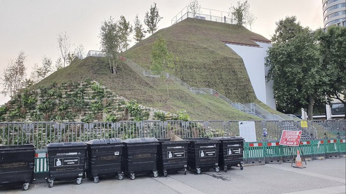 Photo of a man-made grassy mound with a row of wheelie bins in front of it.