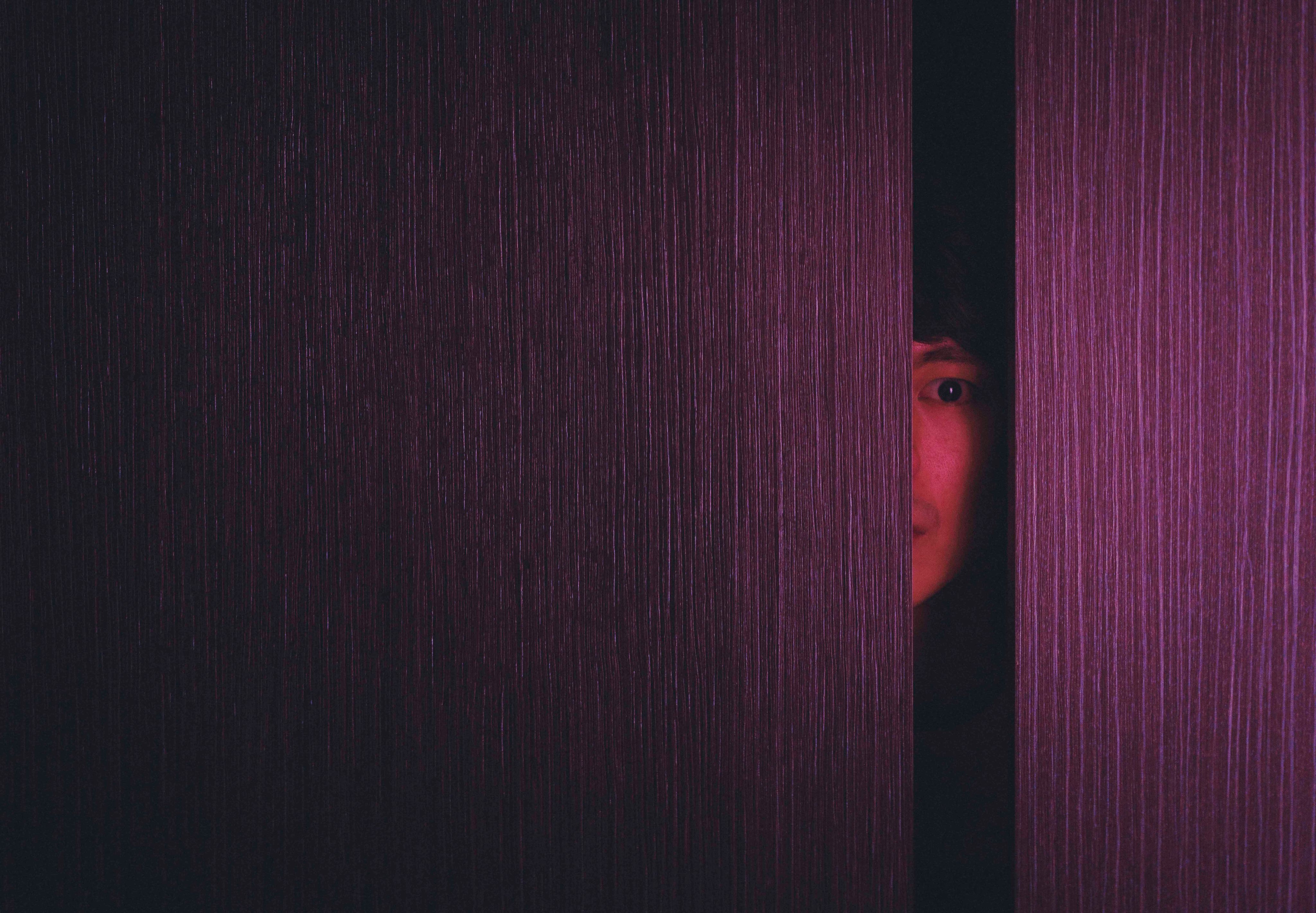 A sliver of Austin Lin's face peers through a door into a dark room with soft purple light. 