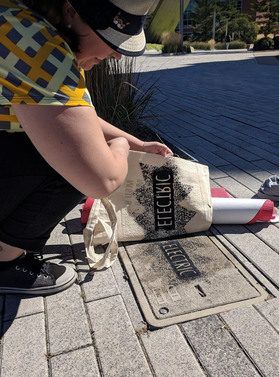 A person is outside, crouched over a utility cover on the ground. The cover reads ELECTRIC and is covered in black ink. The person is pulling a print, lifting a tote bag off of the utility cover, and the ELECTRIC black print is reflected on the tote bag.