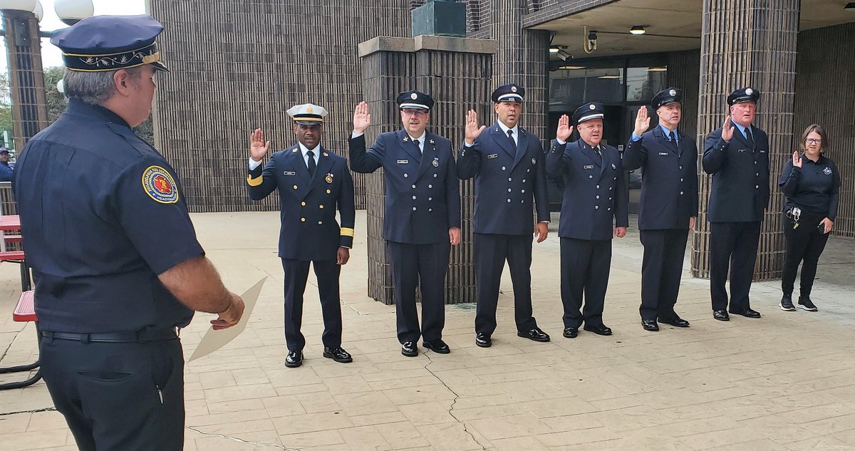 six men in dress uniforms and one woman stand outdoors on patio with right hands raised and looking toward man holding piece of paper