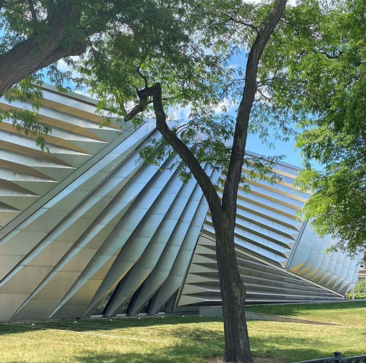 The MSU Broad Art Museum exterior. Reflected in the steel architecture is the blue sky and green grass surrounding it. A green tree is in the foreground of the photo.