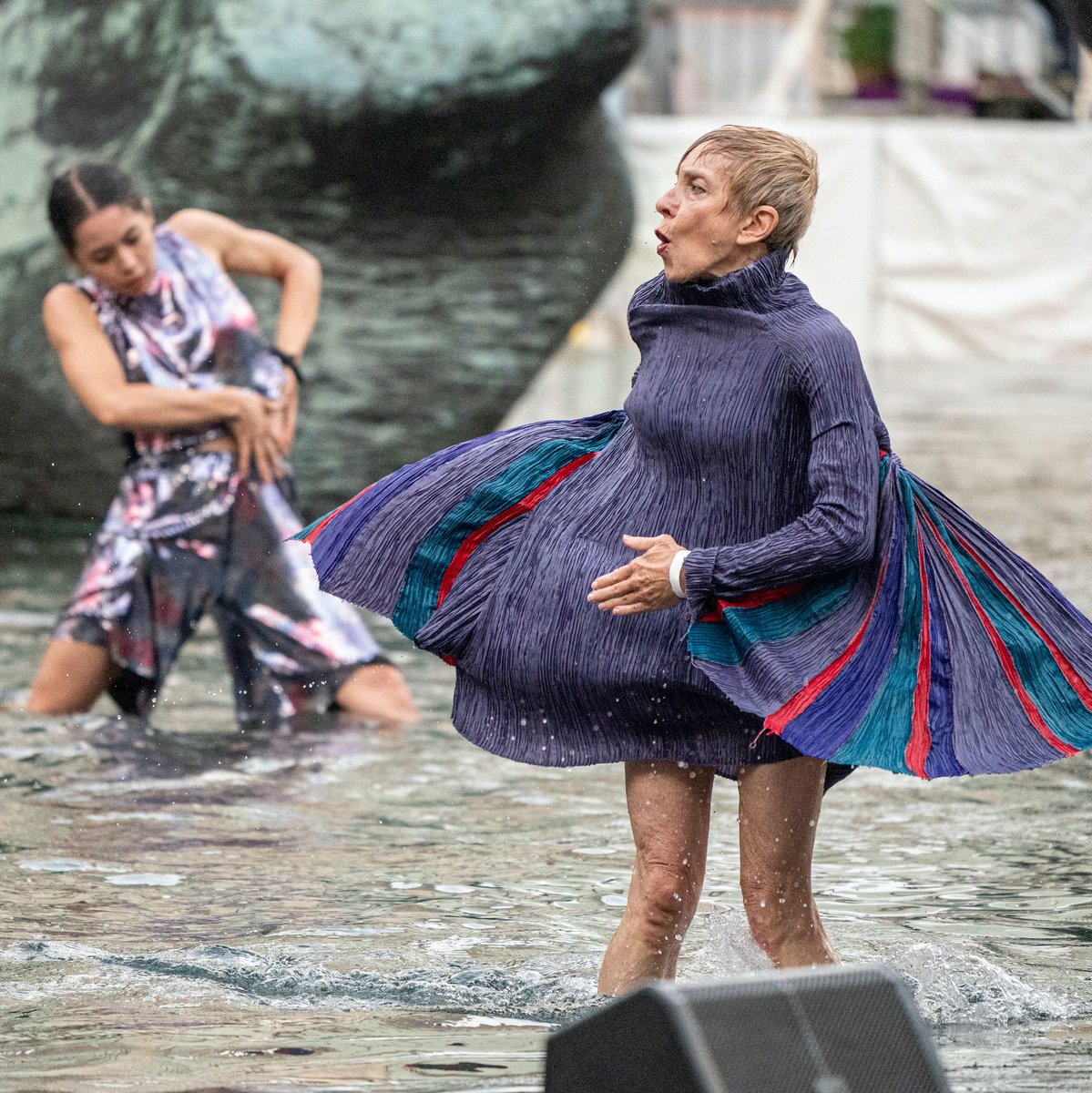 A close up image of a white female cast member dancing in the Paul Milstein Reflection Pool. The woman's dress is purple with green, blue, and red stripes. Other cast members can be seen in the background.