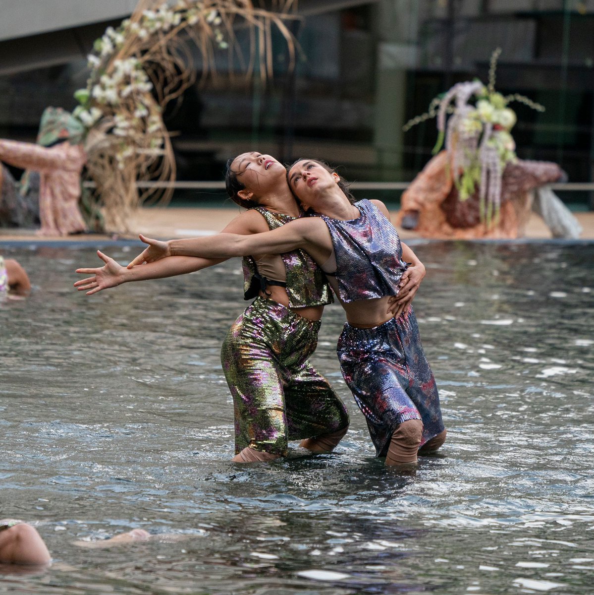 An image of two female cast members dressed in yellow and purple outfits perform together in the Paul Milstein Reflection Pool. Sculptures near the pool can be seen in the background.
