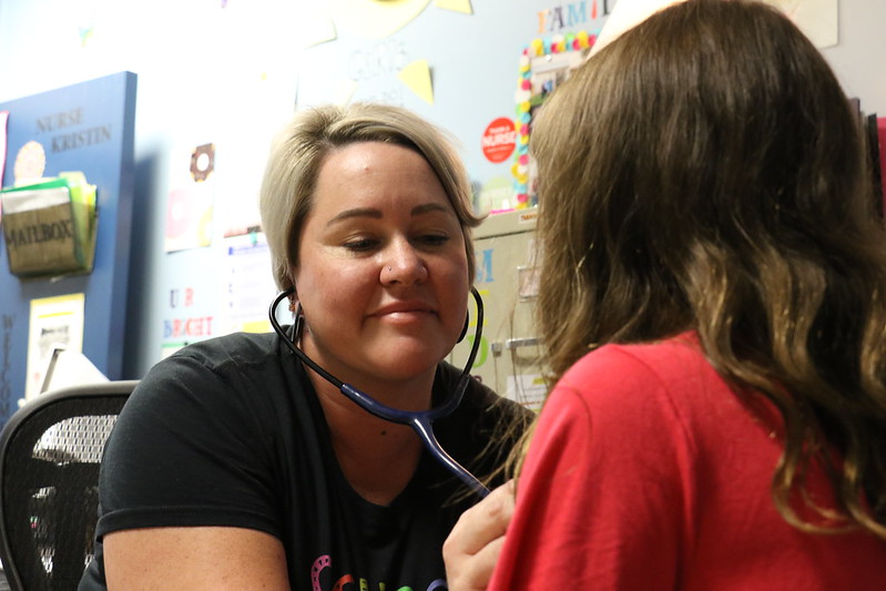 Nurse Kristin sitting in her office, smiling while using a stethoscope to listen to a child's heart.