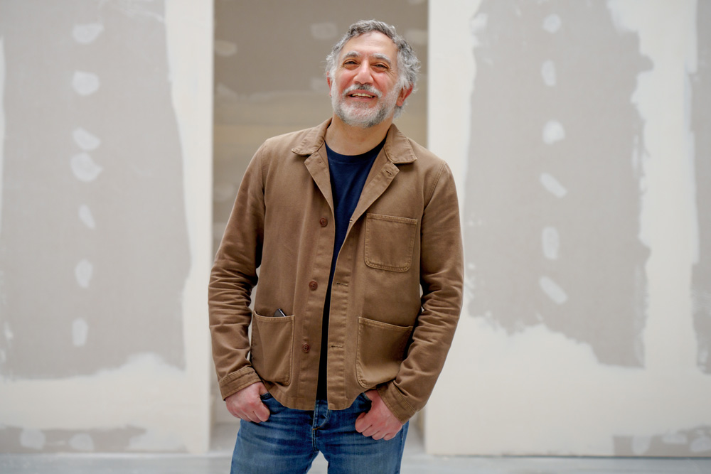 Hashim Sarkis is standing in front of an unpainted wall. His thumbs are hooked in his jean pockets and he has a big smile on his face.