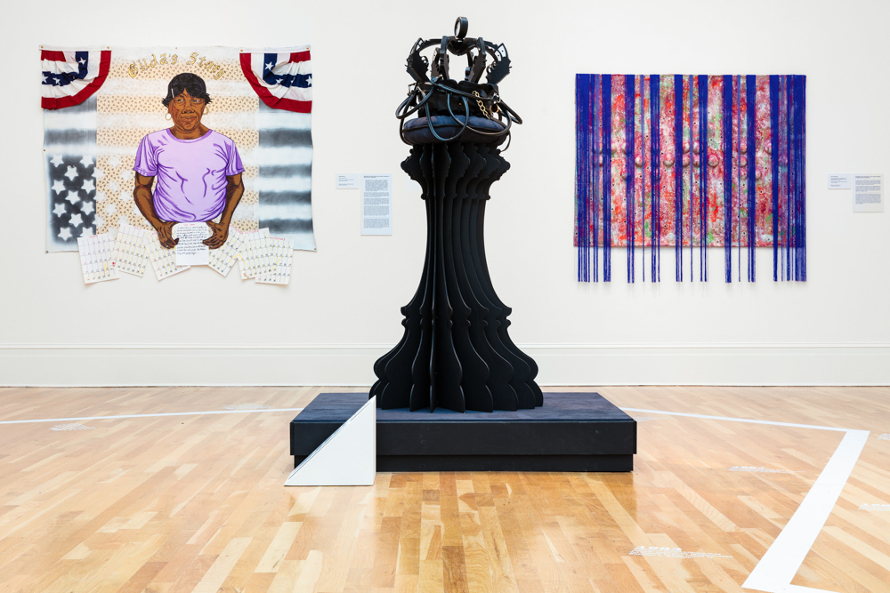 A gallery shot of the Per(Sister) exhibition. A large black sculpture is in the center of the gallery. A thick, white line divides the gallery floor. Artwork hangs on the wall.
