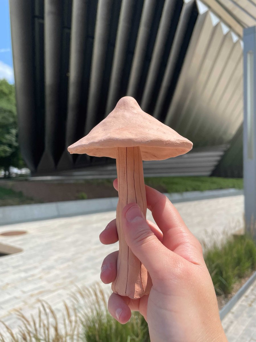 A small, handmade clay mushroom is held by a hand in front of the MSU Broad Art Museum’s architecture.