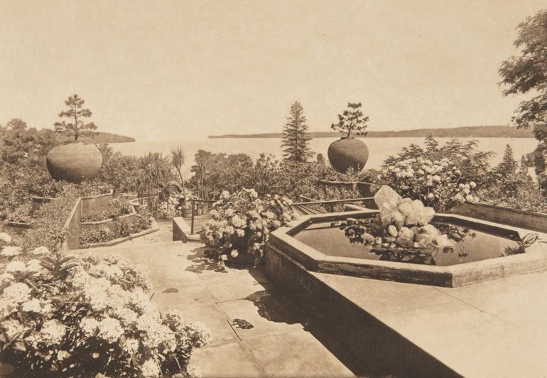 Sepia image of a garden designed by Louis C. Tiffany. The garden is on multiple levels with a pond in the foreground, two trees in huge round pots, and palm trees at the lowest level in the distance. Beyond the garden is an expansive view of the water surrounding Long Island.