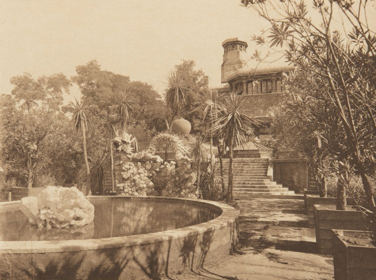 Sepia image of a garden and home designed by Louis C. Tiffany. In the foreground is a constructed pond with a large glass sculpture in the middle. The garden surrounding the pond has palm trees, other sculptures, and steps leading up to the house in the background.
