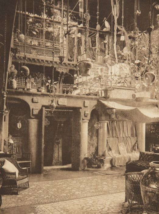 Sepia image of the interior of a house designed by Louis C. Tiffany. The room is large, of double height, and decorated by a multitude of objects, some hanging from the ceiling.