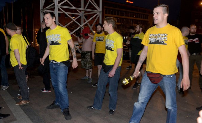 Nazis mit gelben "Weg mit dem NWDO-Verbot" stehen vor dem Rathausplatz. Lukas Bals hält eine Sektflasche am Flaschenhals fest (so als könne man damit gut zuschlagen)